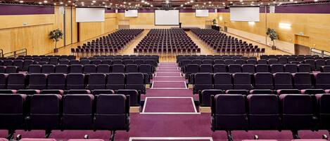 Un gran auditorio con filas de sillas moradas, suelo de madera y un escenario con una pantalla grande al frente.
