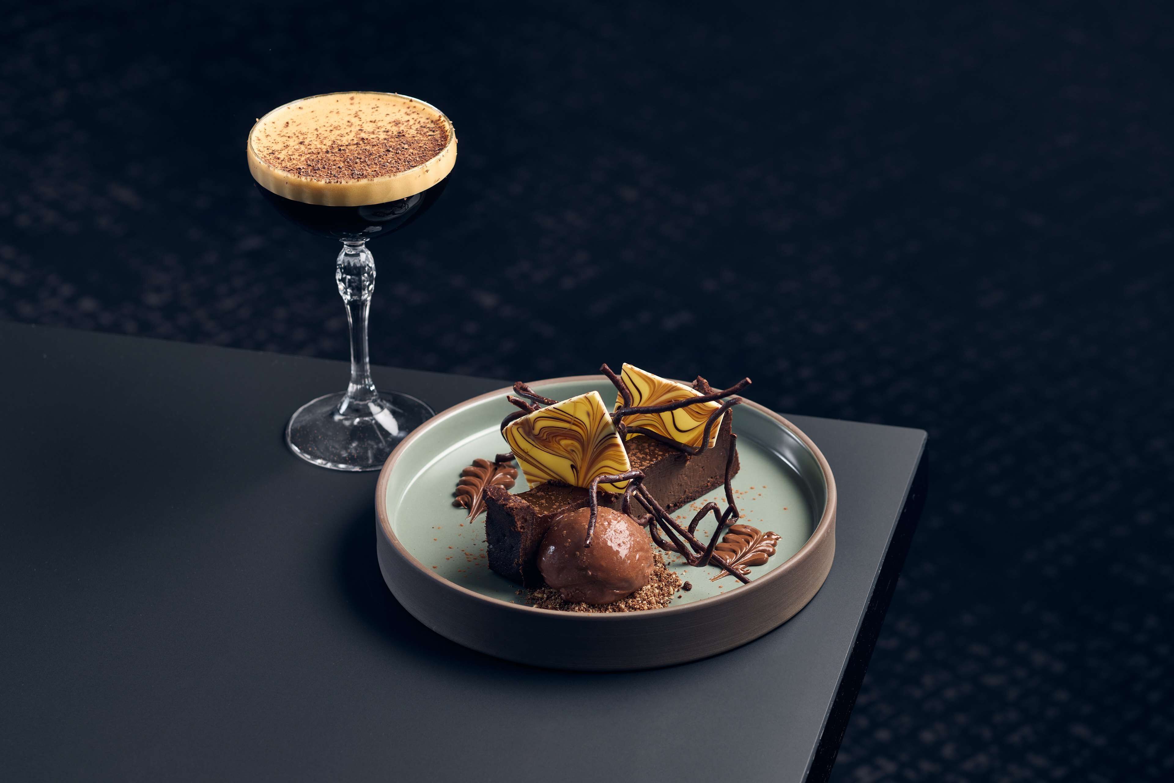 Un plato de postre con un dulce de chocolate y un vaso de bebida oscura sobre una mesa oscura.