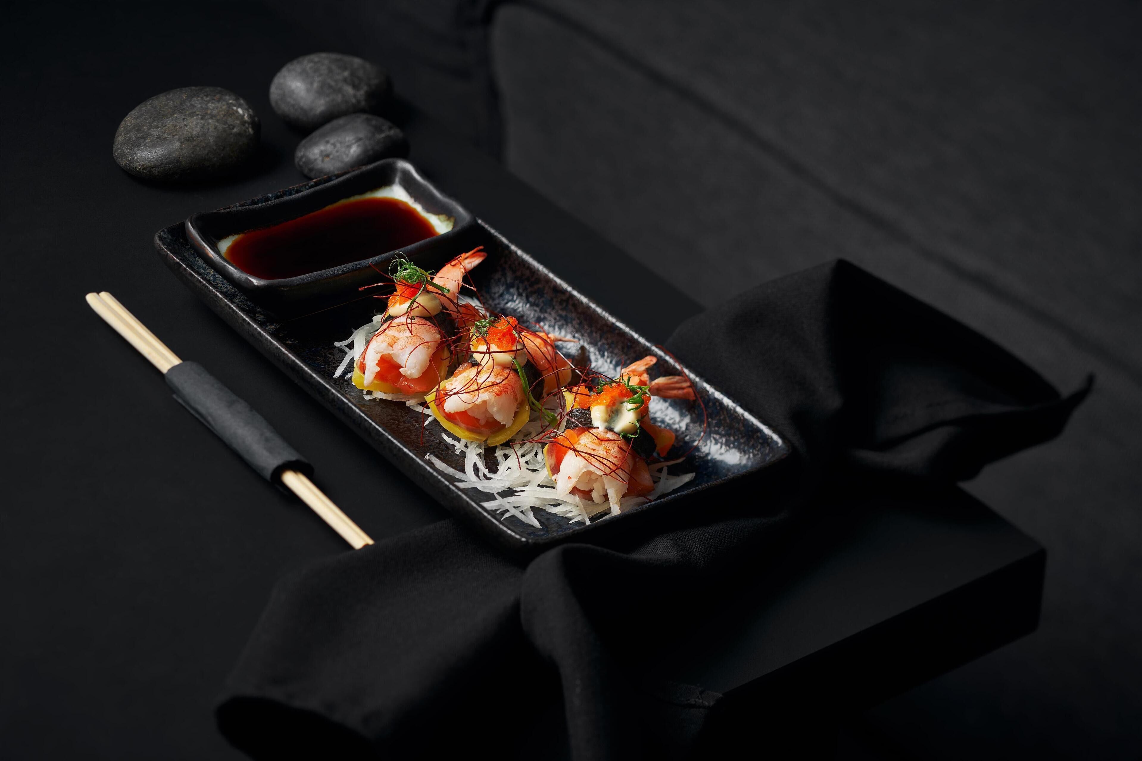 A dish of shrimp on a black plate with chopsticks and a small bowl of sauce.