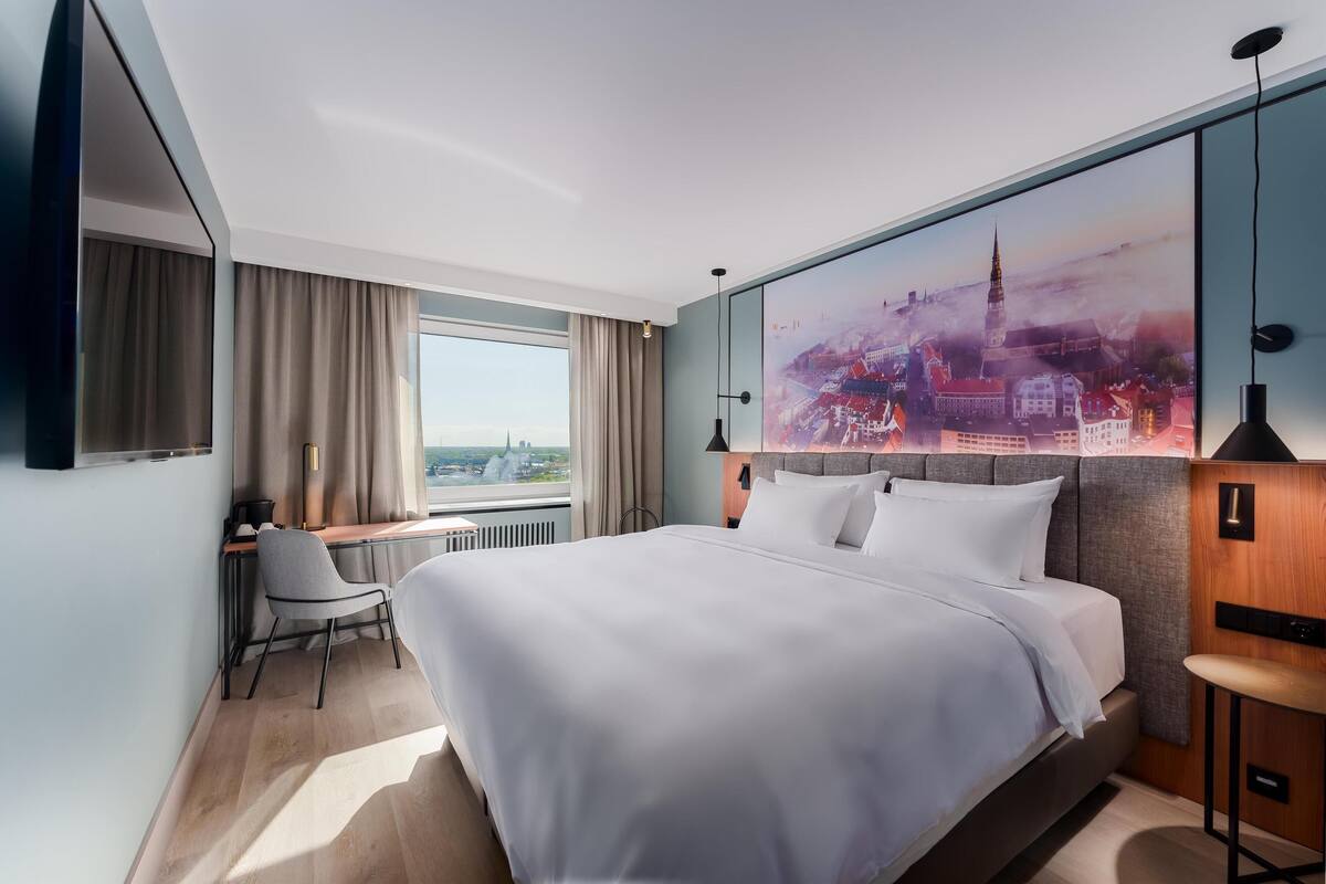 Una habitación de hotel moderna con una cama grande, un escritorio con silla, un ventanal con cortinas y un mural con vista a la ciudad en la pared.