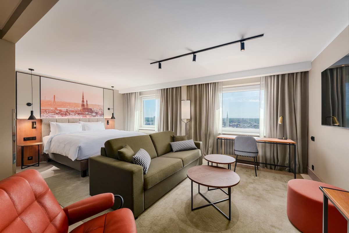 Habitación de hotel con cama, sofá, escritorio y silla. Amplia ventana con cortinas, vista panorámica de la ciudad y un mural con el horizonte urbano.