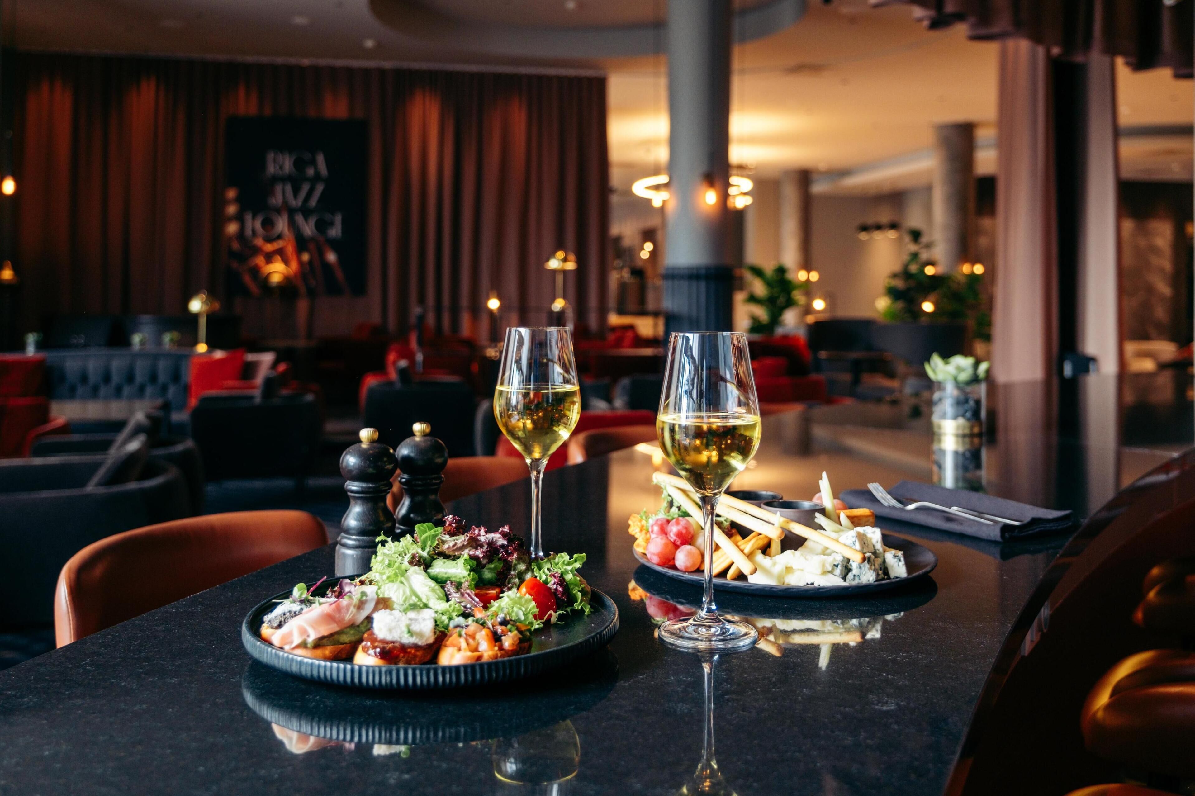 A restaurant setting with a table set for two, featuring a salad and a plate of cheese and crackers, accompanied by two glasses of white wine.