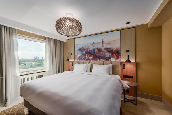 A hotel room with a large bed, a cityscape painting on the wall, a view of the city through the window, and a modern chandelier.