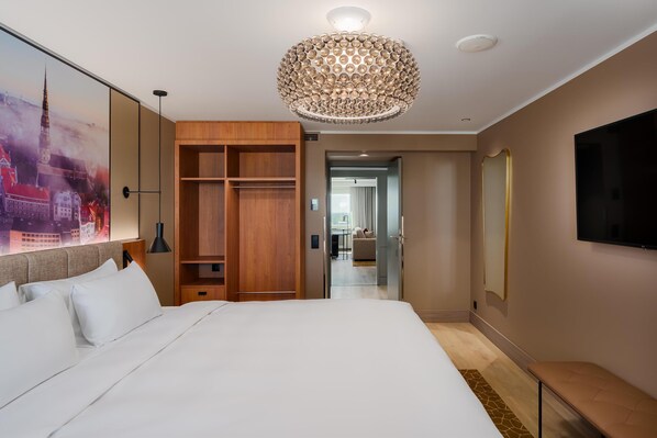 A hotel room with a large bed, wooden wardrobe, a chandelier, a flat-screen TV, and a cityscape painting.