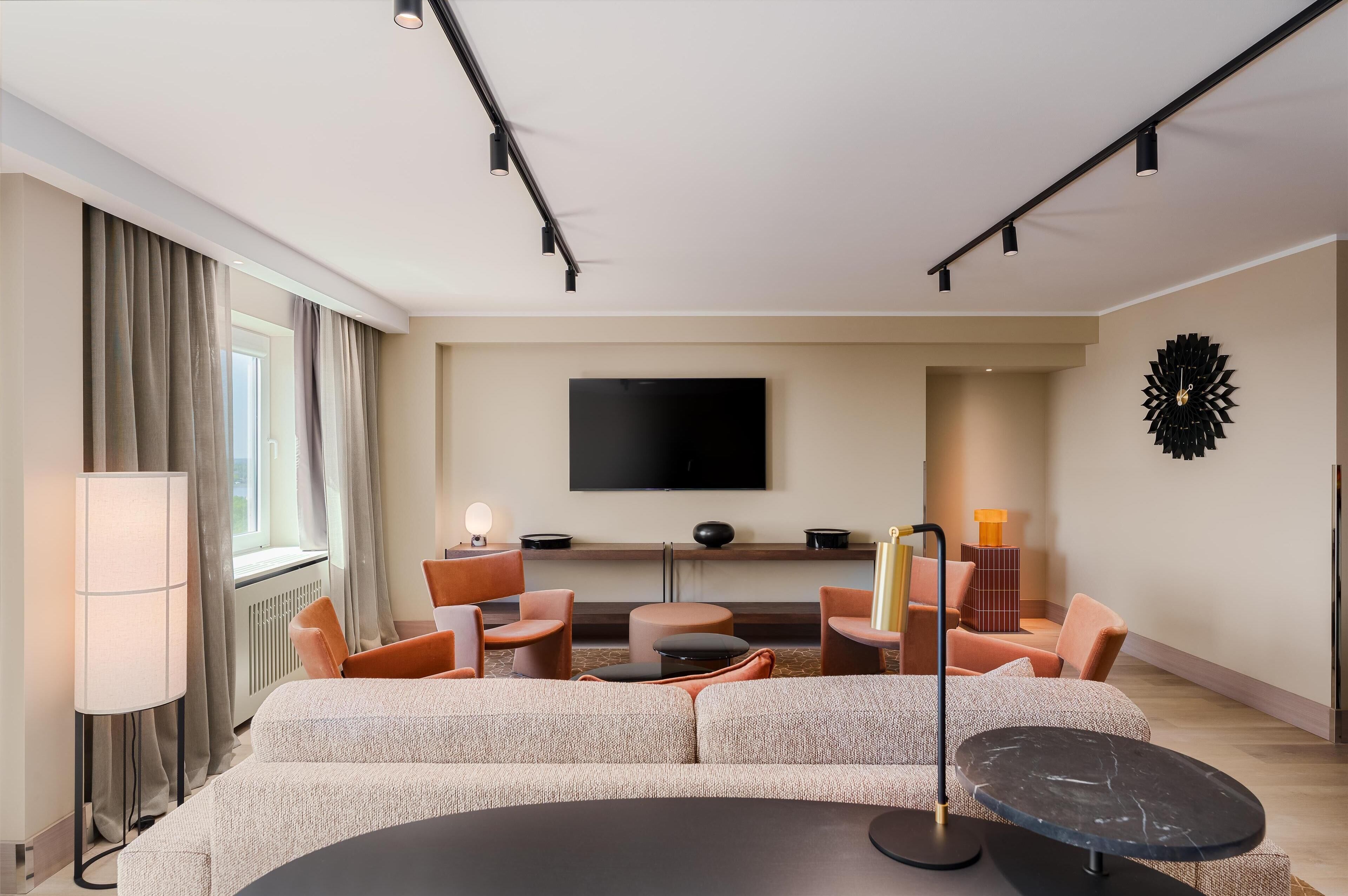 A modern hotel room with a large sofa, a flat-screen TV mounted on the wall, a dining table, and a unique wall decoration.