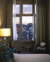 Ein Hotelzimmer mit Bett, Stuhl, kleinem Tisch mit Lampe und Fenster mit Vorhängen.