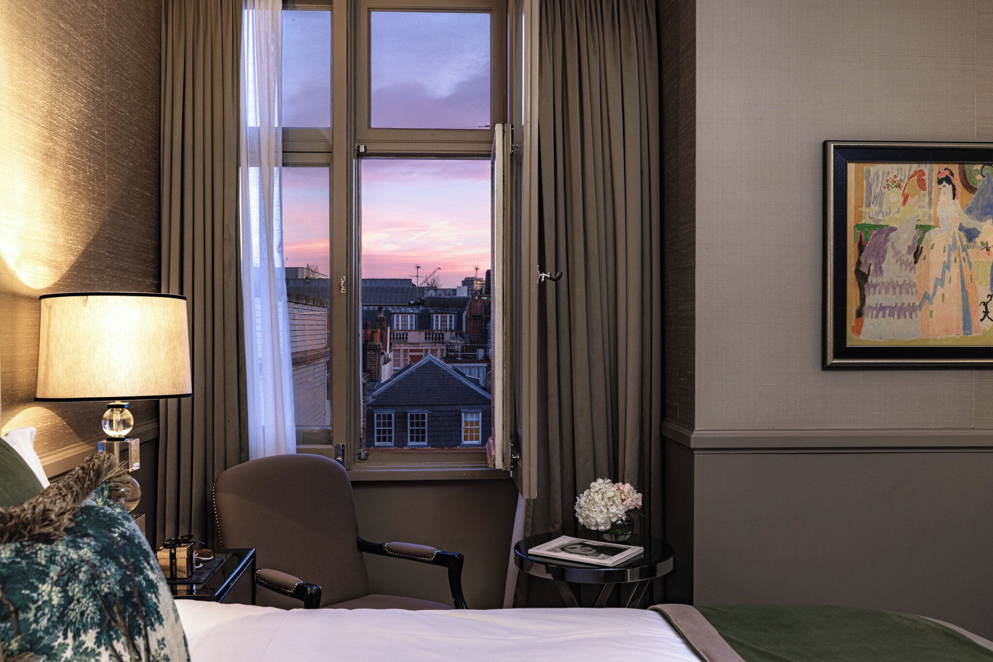 A hotel room with a bed, a chair, a table with a lamp, a window with curtains, and a painting on the wall.