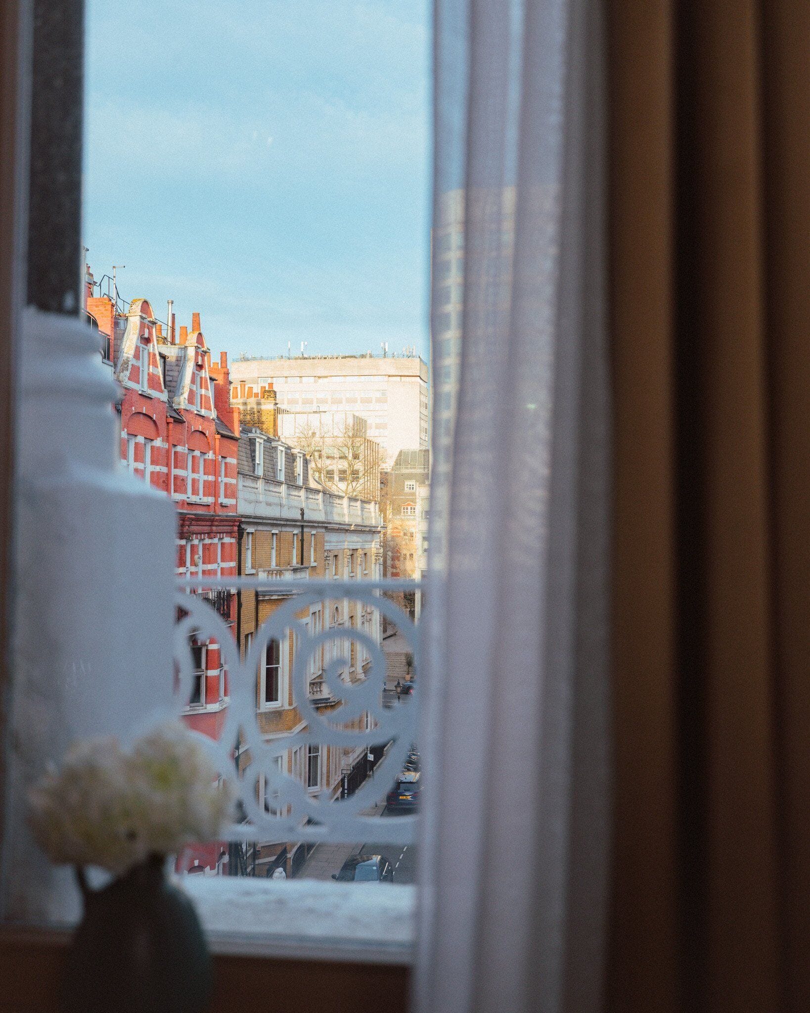 A view through a window with a curtain, featuring a street scene with buildings and a white metal railing.