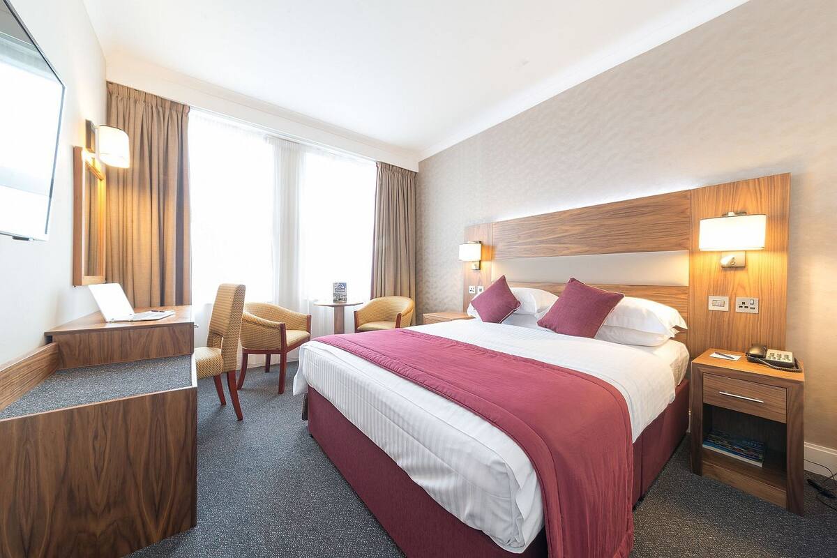 A hotel room with a large bed, a desk with a laptop, a chair, a bedside table with a lamp, and a small table with a lamp.