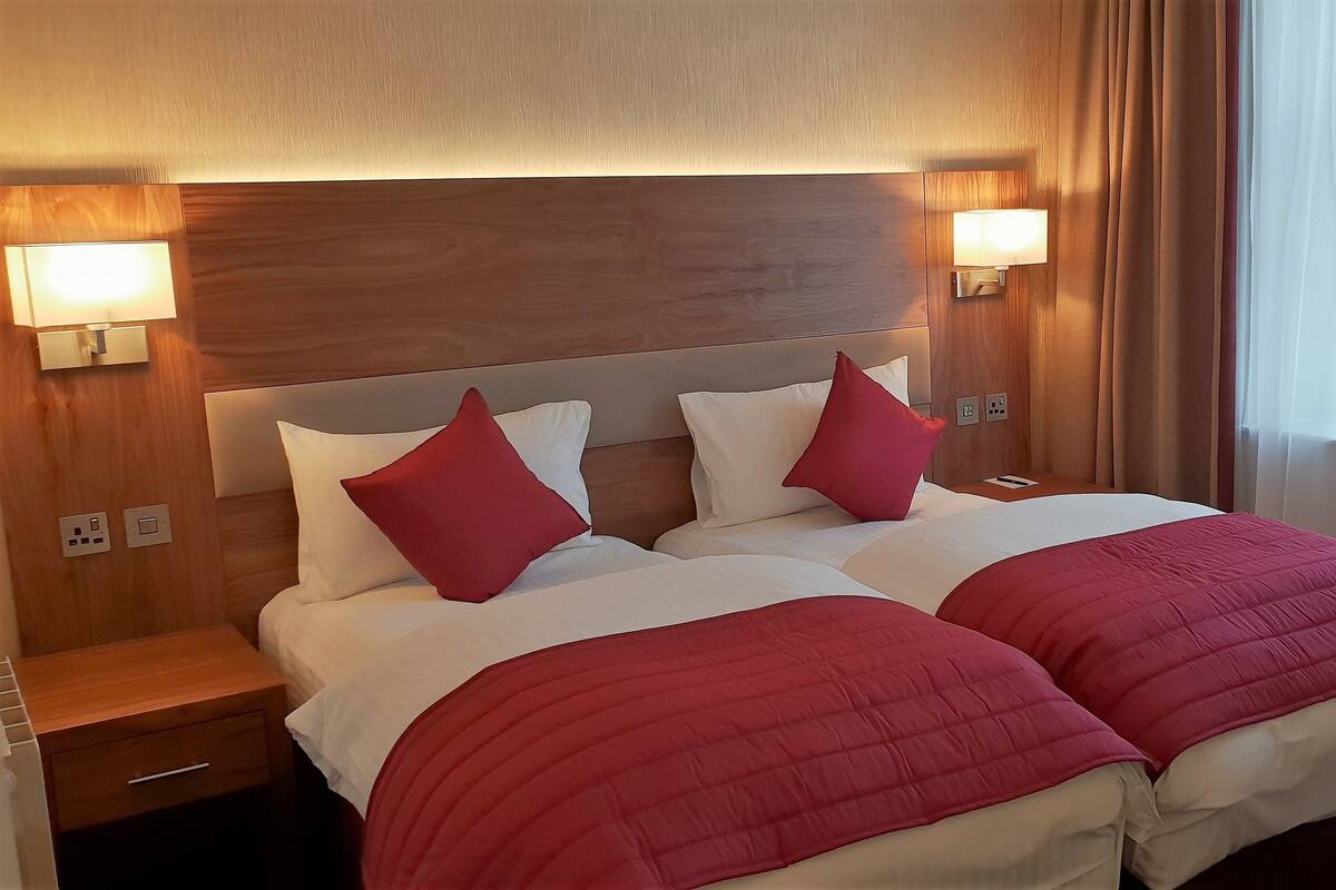 A hotel room with a wooden headboard, two bedside tables, a bed with white and red bedding, and two wall-mounted lamps.