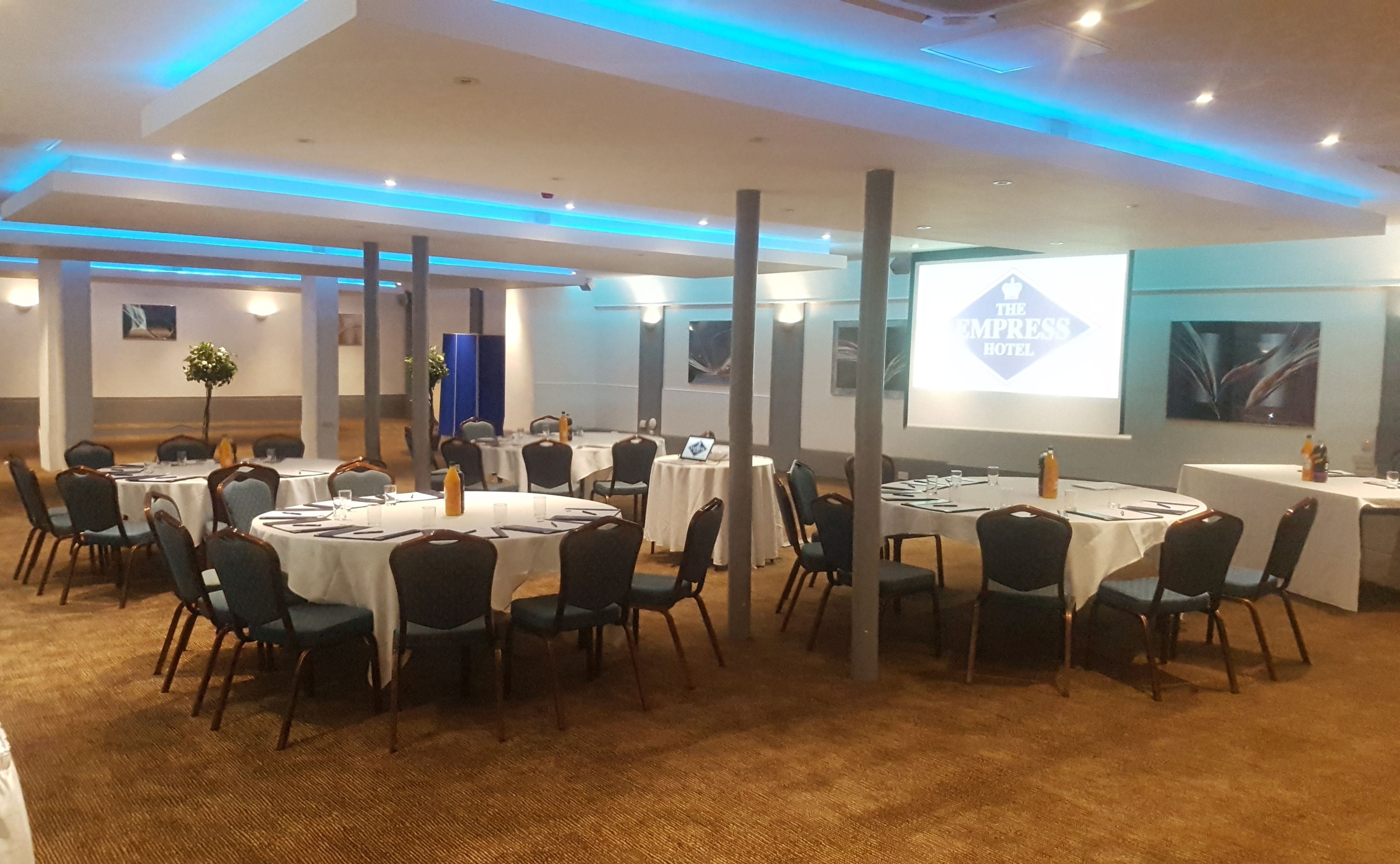 A conference room with multiple tables and chairs, a projector screen displaying 'The Empress Hotel', and a blue-lit ceiling.