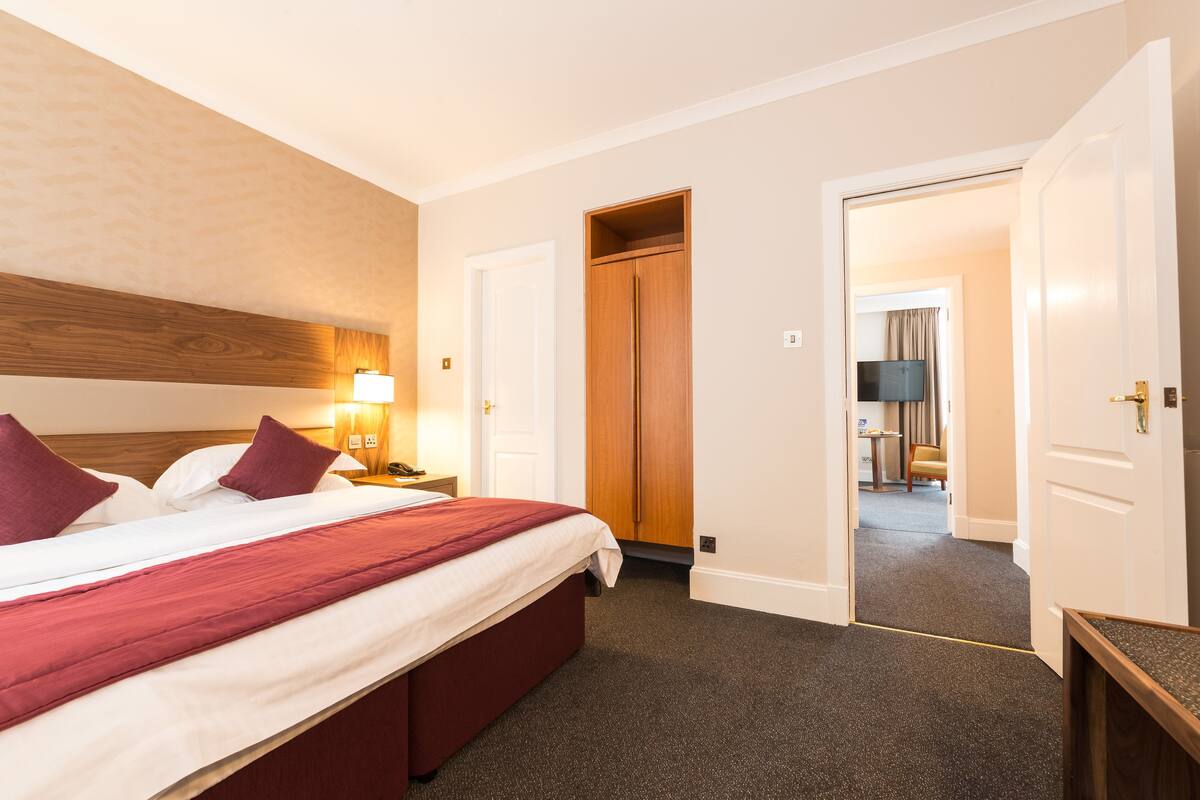 A hotel room with a bed, bedside lamp, wooden headboard, and a door leading to a hallway.