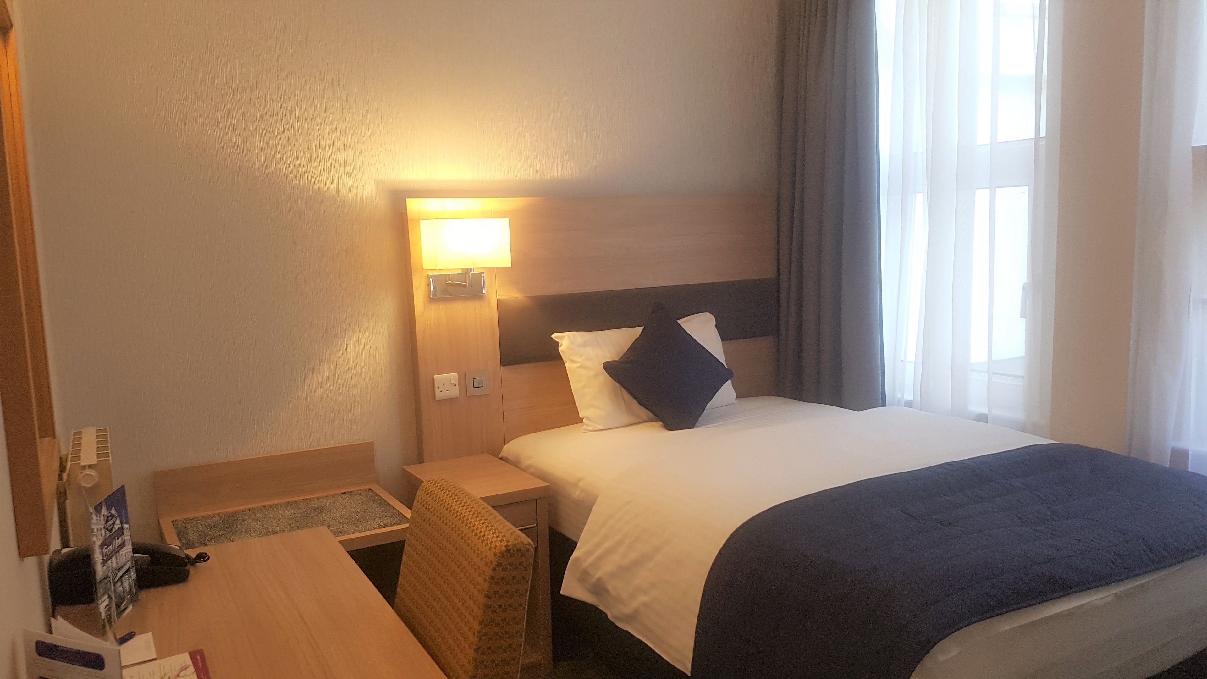 Habitación de hotel con cama, mesita de noche, lámpara y vistas al exterior.