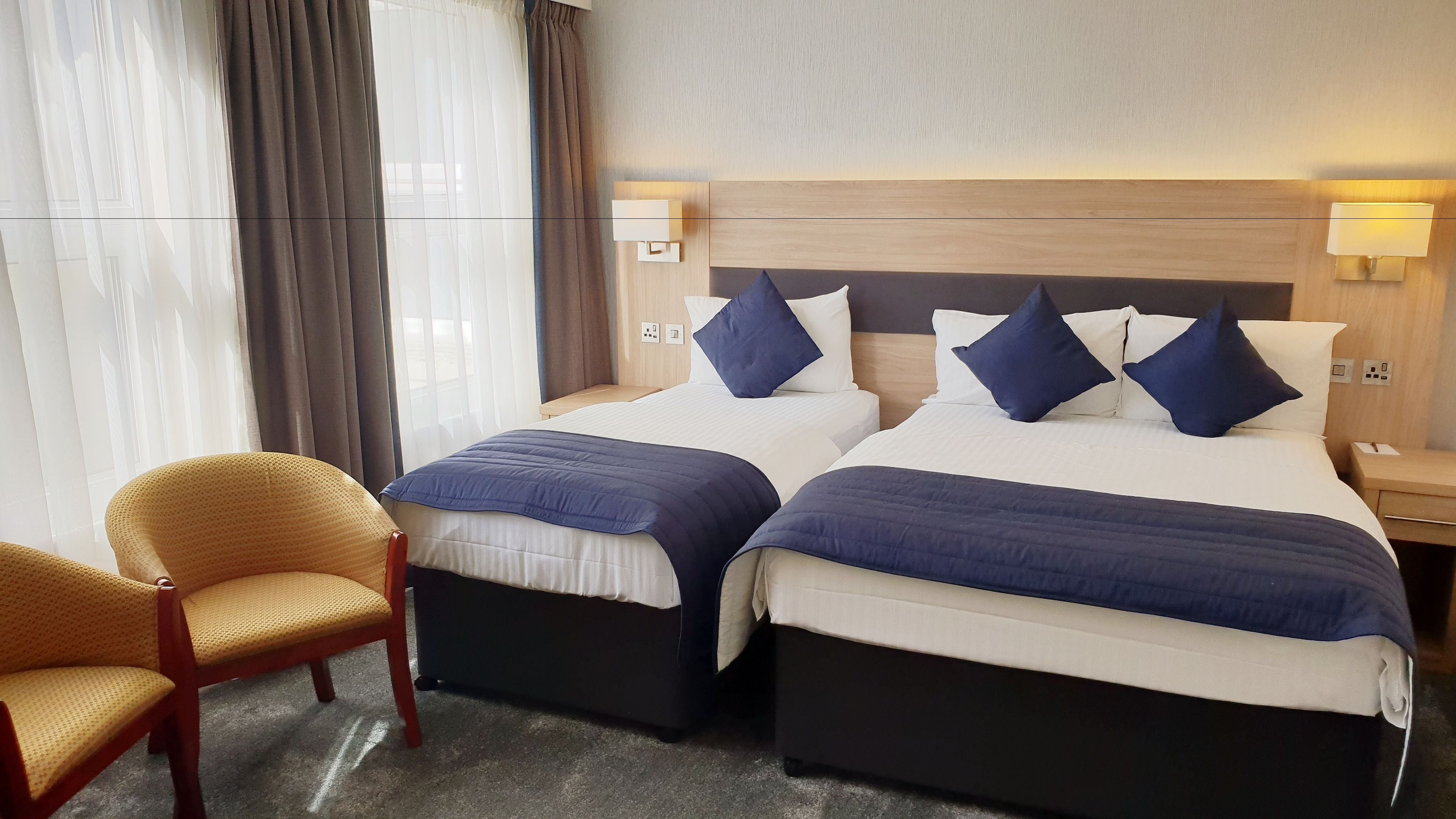 Habitación de hotel con dos camas, cada una con cabecera, una silla y una mesita de noche con lámpara.
