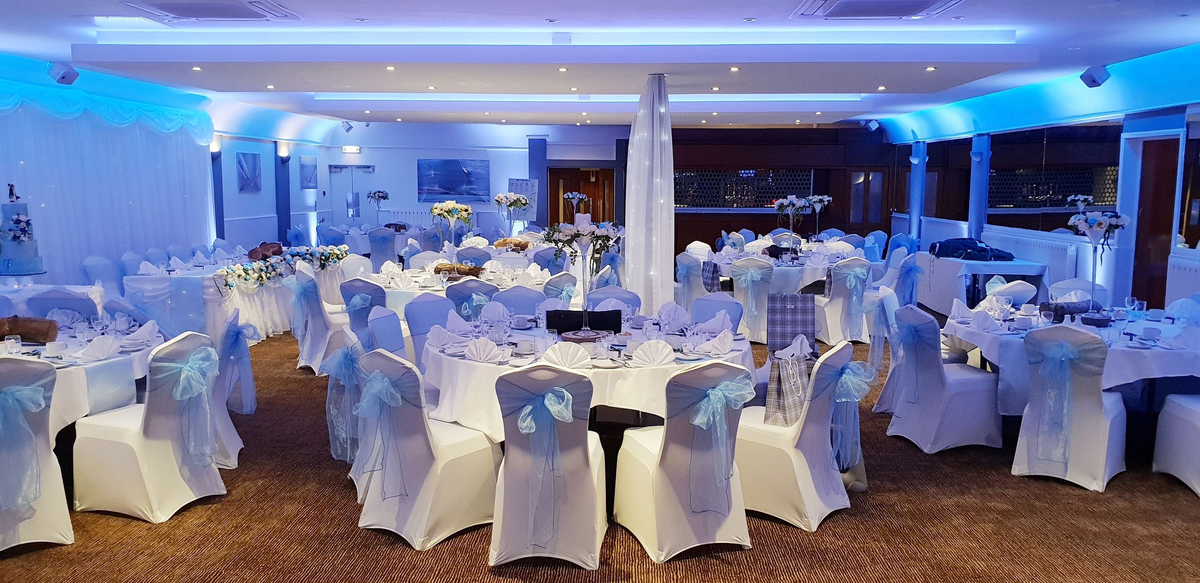 Un salón de banquetes con mesas redondas, manteles blancos y sillas adornadas con cintas azules.