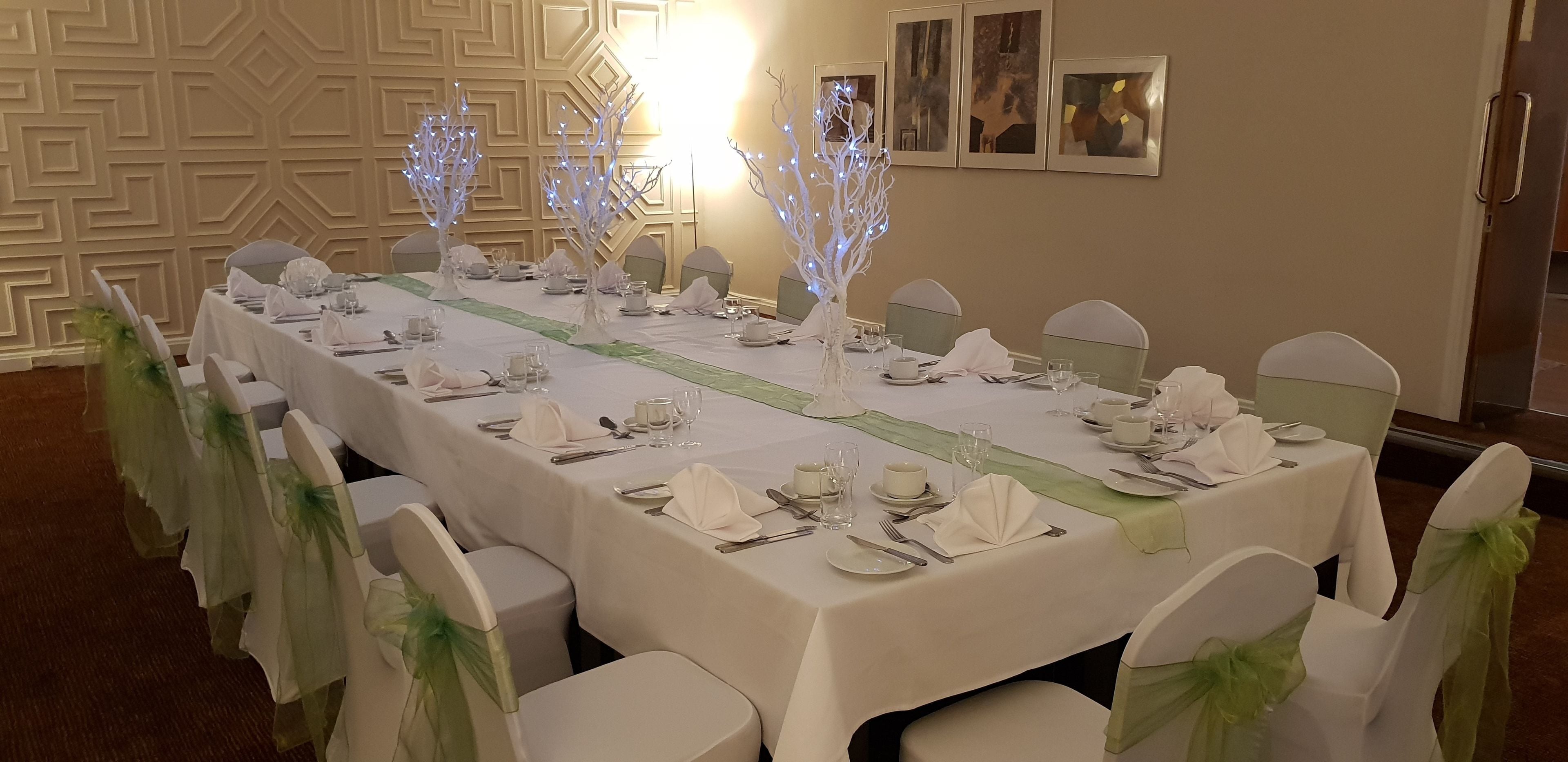 A banquet hall with a long table set for a formal event, featuring white tablecloths, green sashes on chairs, and decorative light trees.