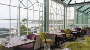 Ein Restaurant mit großen Fenstern, das Meerblick und parkende Autos bietet. Die Tische sind mit weißen Servietten und Weingläsern gedeckt.