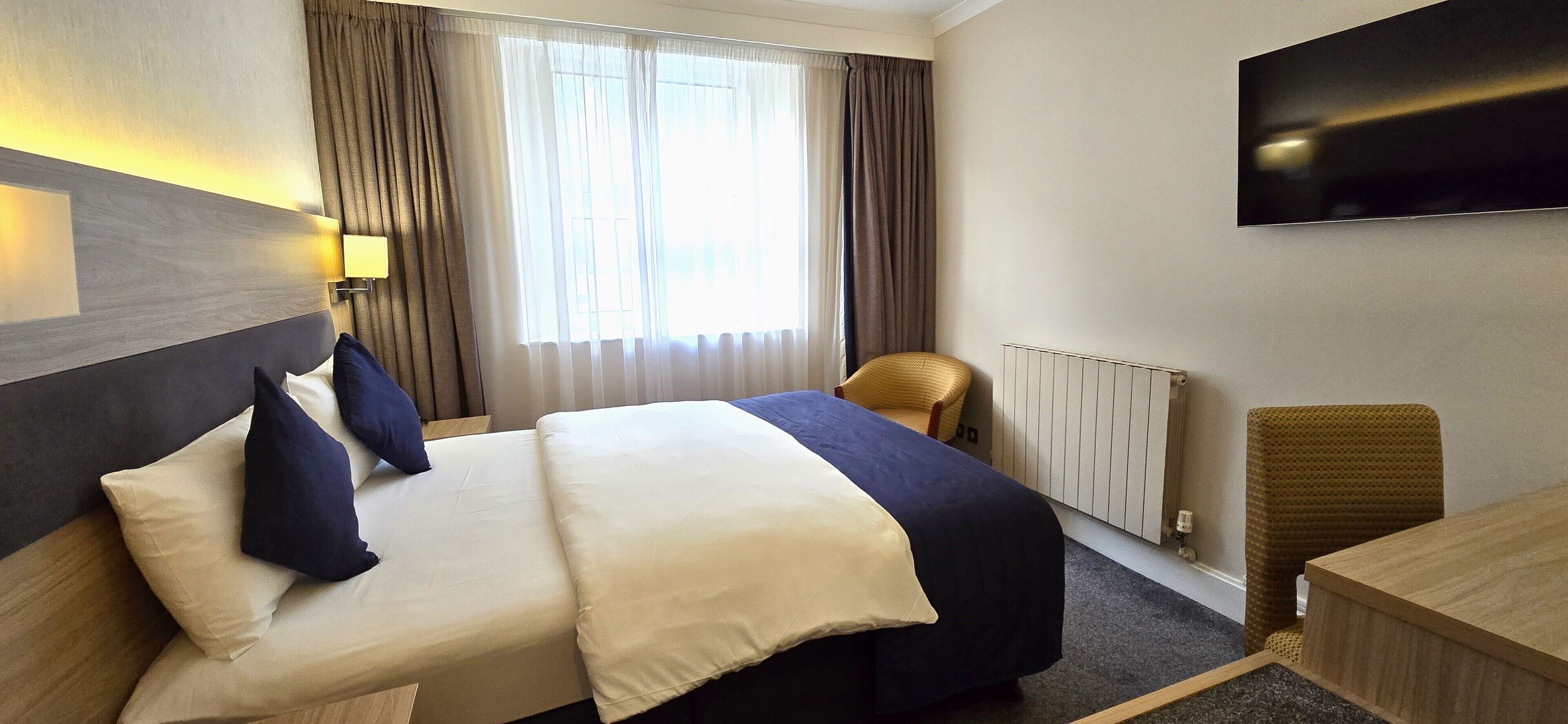 A hotel room with a large bed, a desk, a chair, a TV mounted on the wall, and a window with curtains.