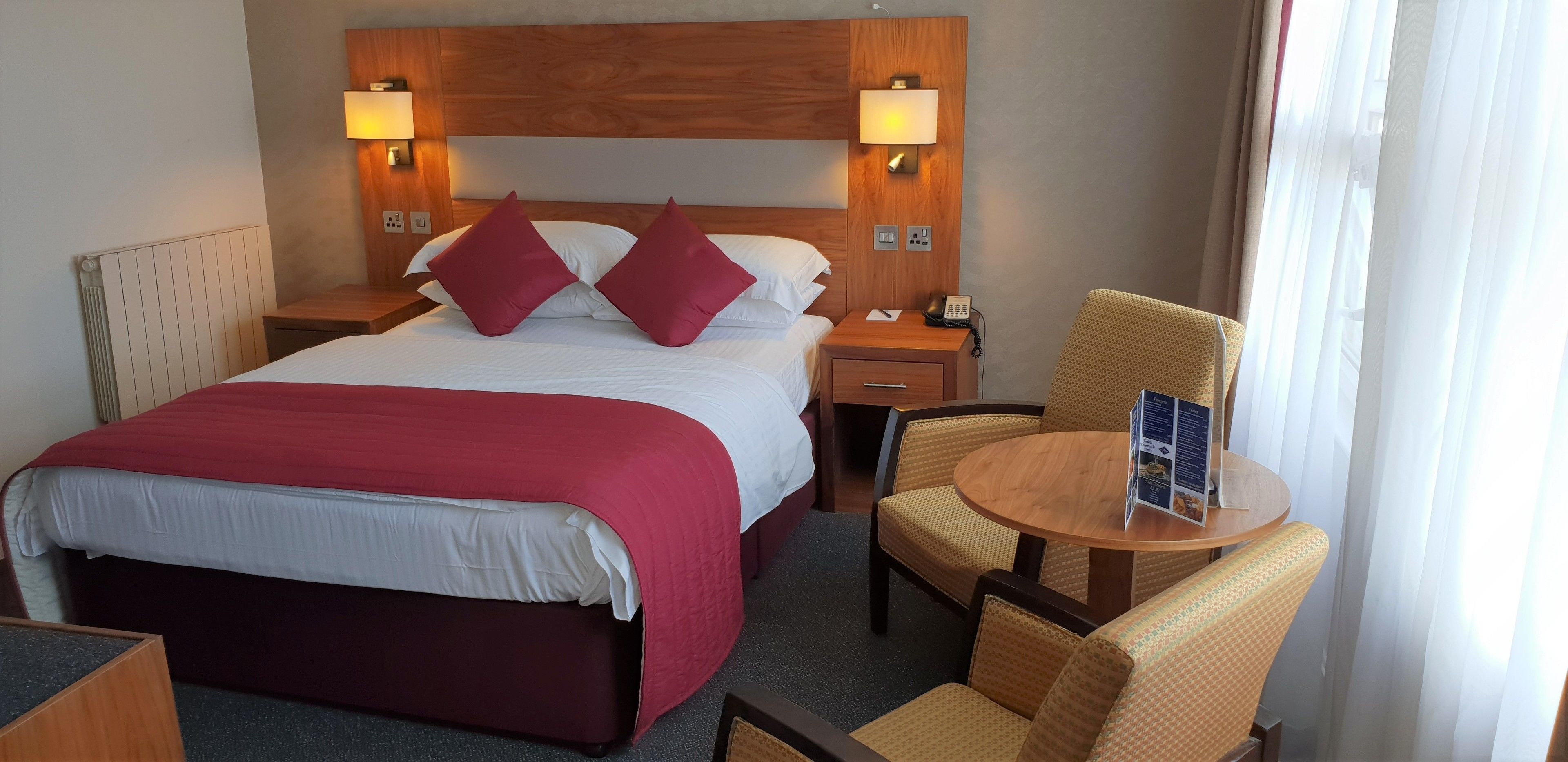 A hotel room with a large bed, two bedside lamps, a wooden headboard, a small table with a menu, and a chair.