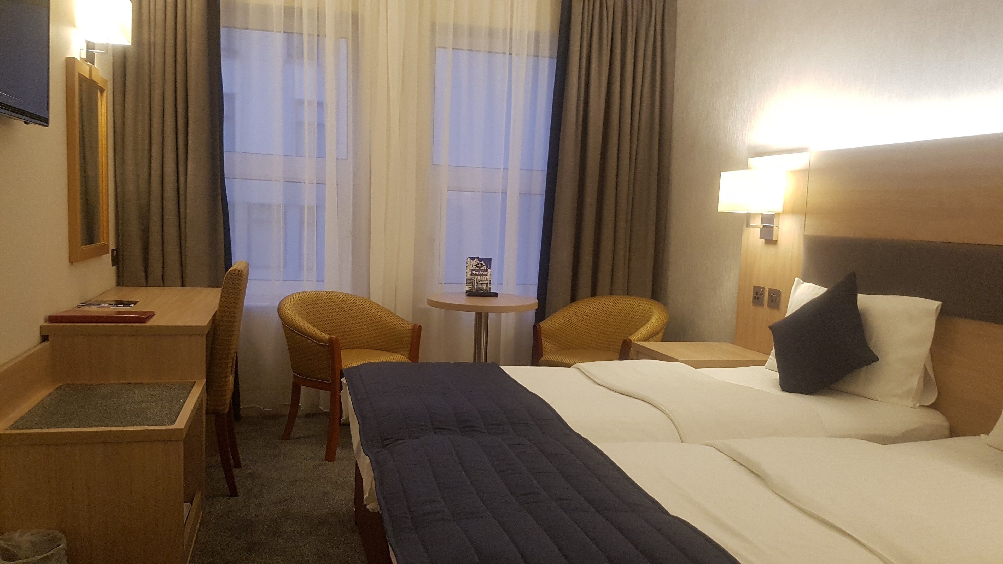 A hotel room with a bed, two chairs, a small table, a desk, and a window with curtains.