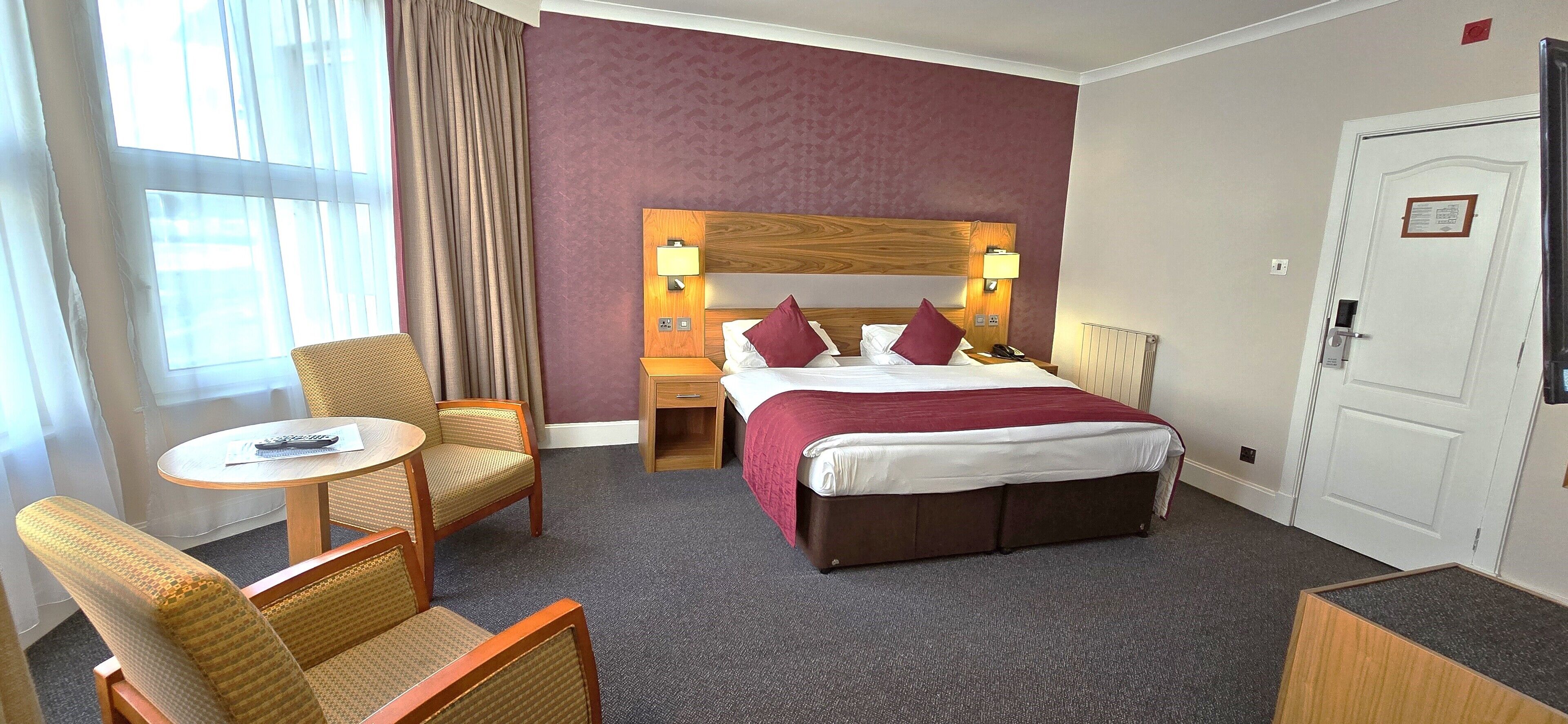 Habitación de hotel con cama, dos mesitas de noche con lámparas, una silla, una pequeña mesa y un ventanal con cortinas.