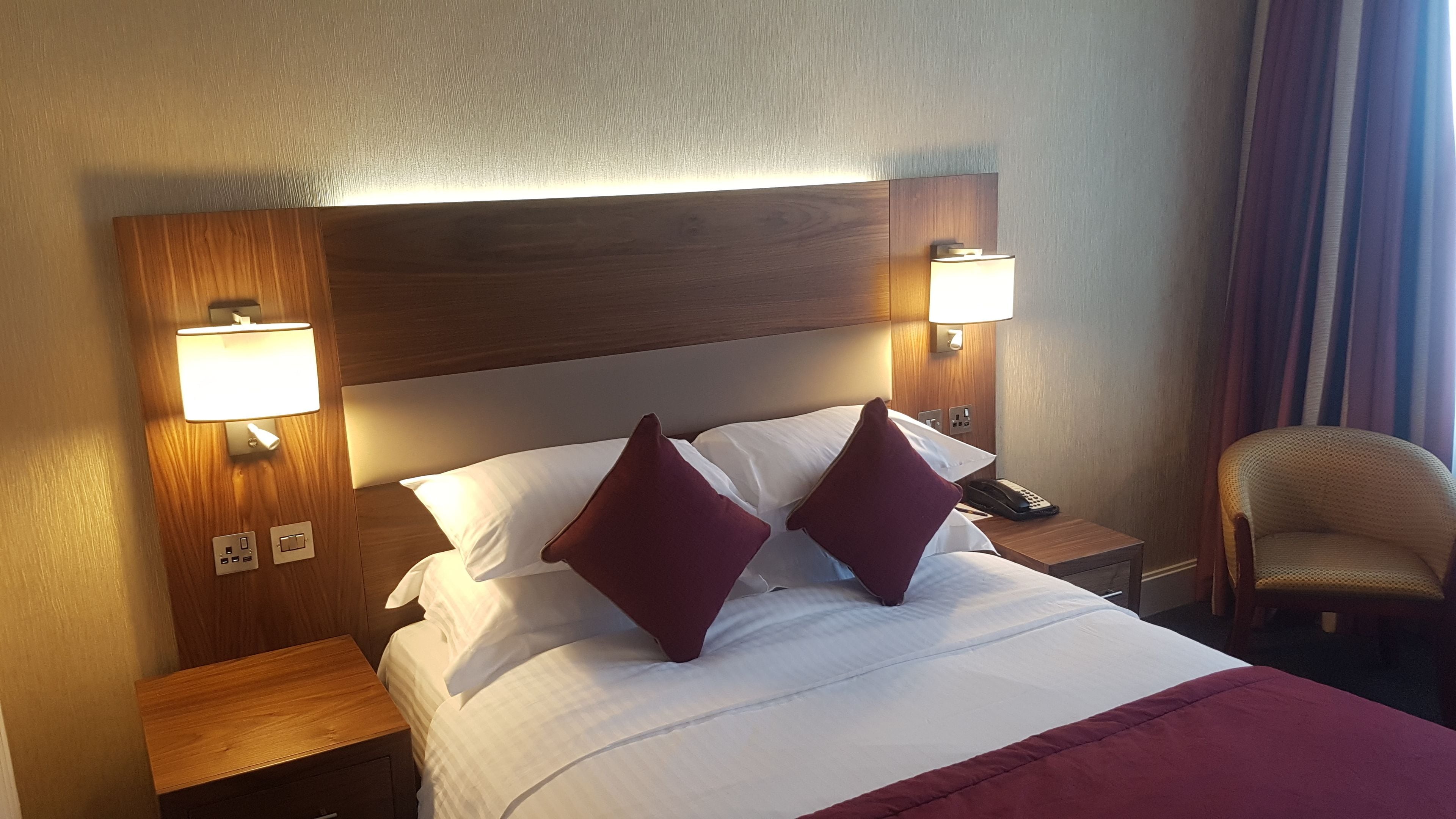 Une chambre d’hôtel avec une tête de lit en bois, deux lampes de chevet, un téléphone et une chaise.