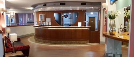 A hotel reception area with a curved wooden reception desk, a mirror, and a TV screen above it.