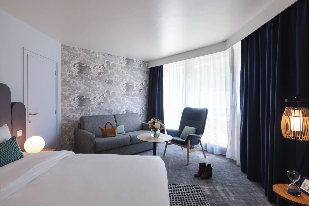 Ein Hotelzimmer mit einem Bett, einem Sofa, einem Sessel, einem kleinen Tisch mit Blumenvase, einer Lampe und einem Fenster mit Vorhängen.