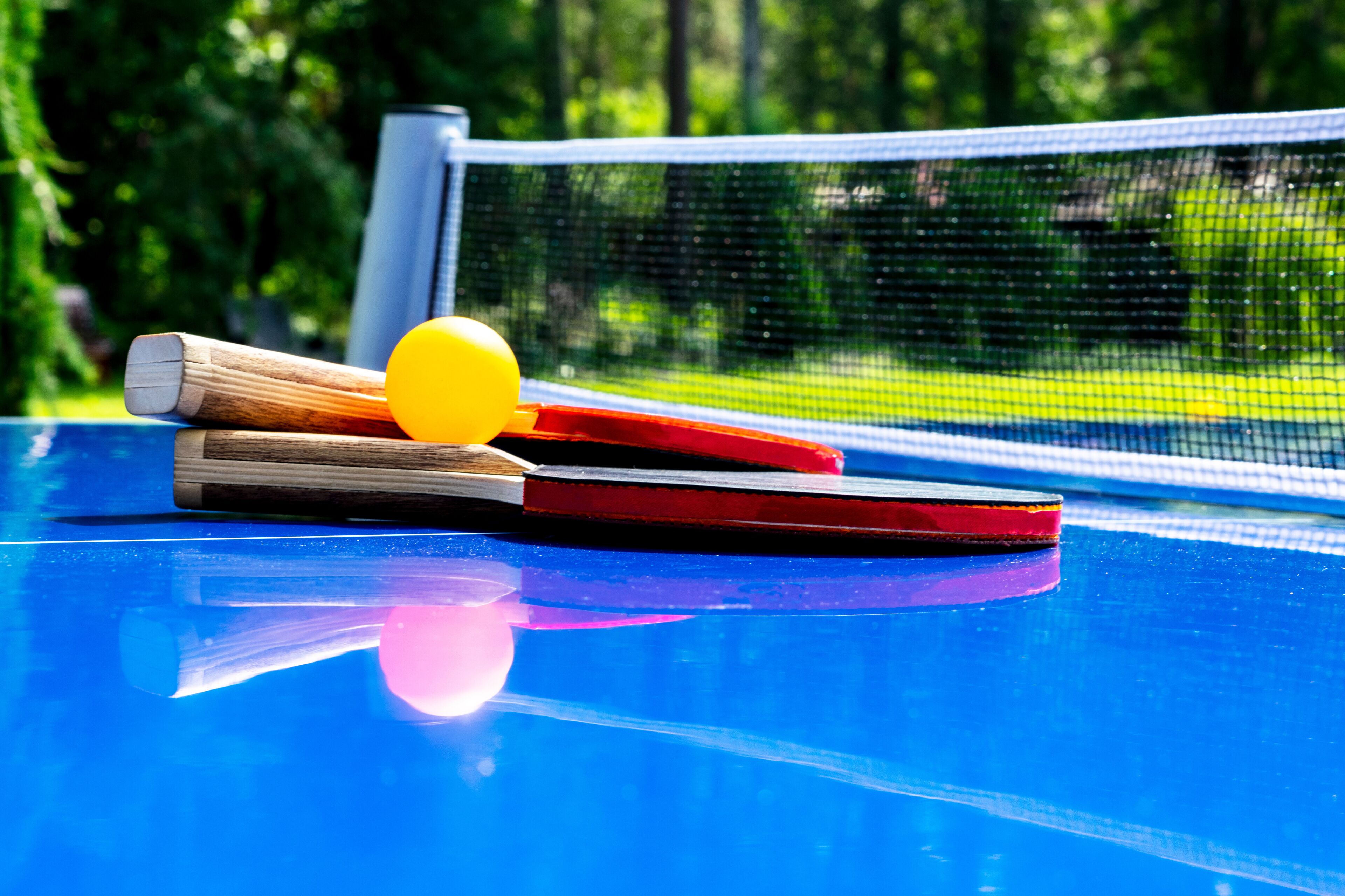 Ein blauer Tischtennis-Tisch mit Netz, einem gelben Ball und Holzschlägern.