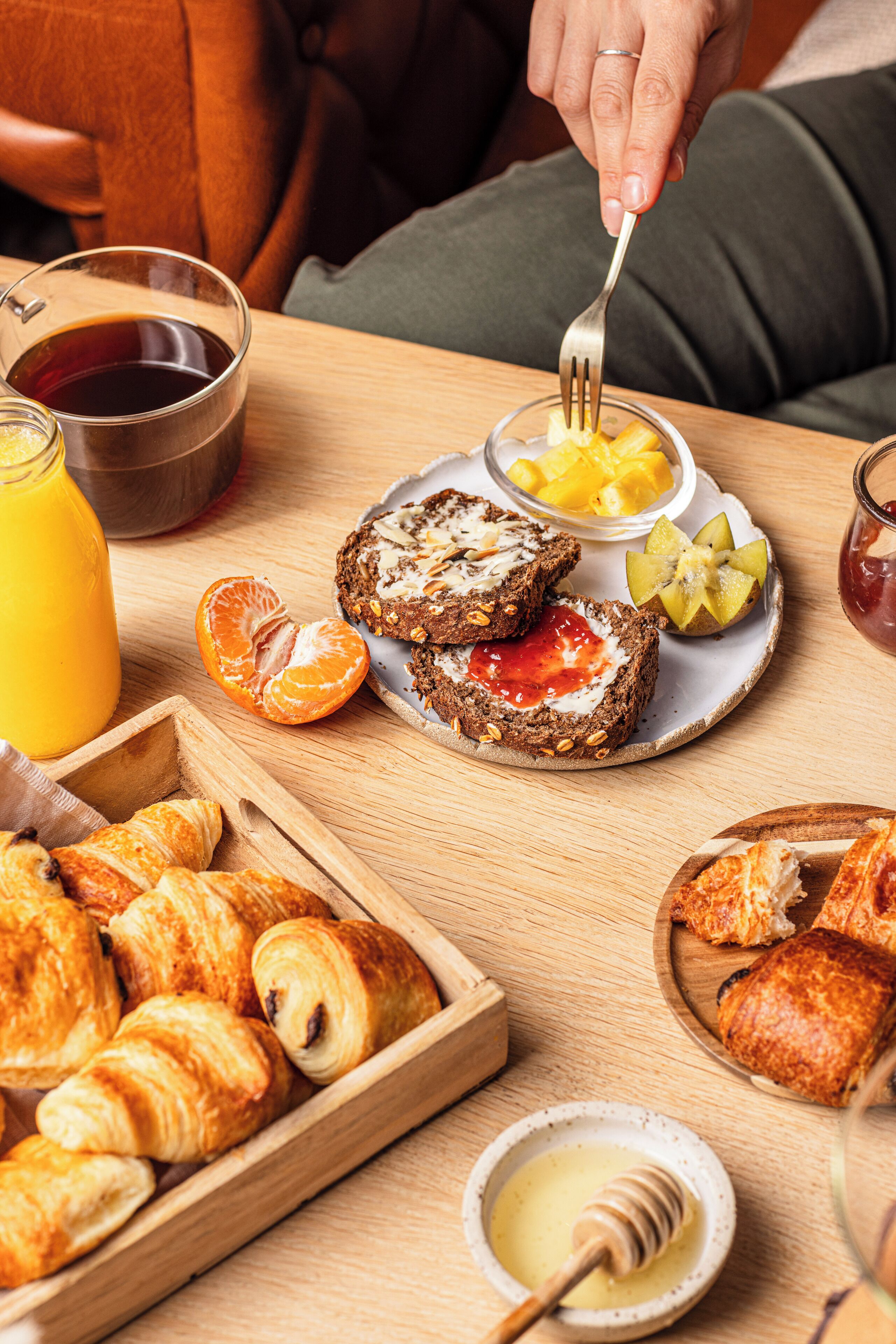 A breakfast setting with croissants, toast with jam and honey, a glass of orange juice, and a bowl of fruit.