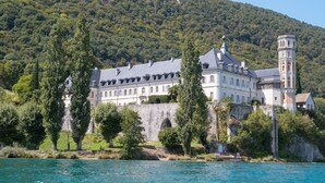A large, historic building with a tower, situated by a body of water, surrounded by lush greenery and a forested hill. - Mercure Hotel & Spa Aix-les-Bains Domaine Marlioz (Aix-les-Bains)