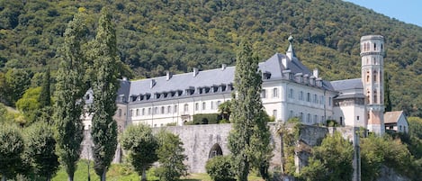 A large, historic building with a tower, situated by a body of water, surrounded by lush greenery and a forested hill.
