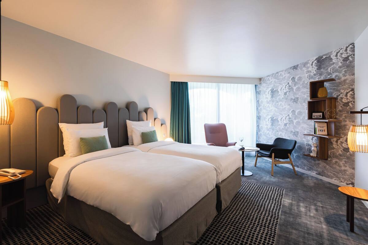 Ein Hotelzimmer mit zwei Betten, einem Schreibtisch, einem Sessel und einem Bücherregal, mit einer Wand mit Muster.