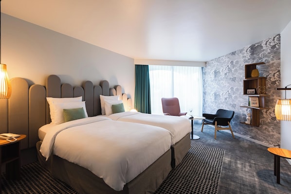 A hotel room with two beds, a desk, a chair, a bookshelf, and a patterned wall.