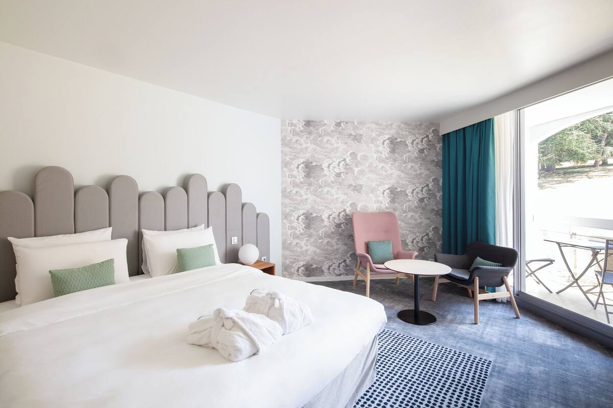 Ein Hotelzimmer mit Bett, kleinem Tisch, zwei Stühlen und Blick ins Freie.