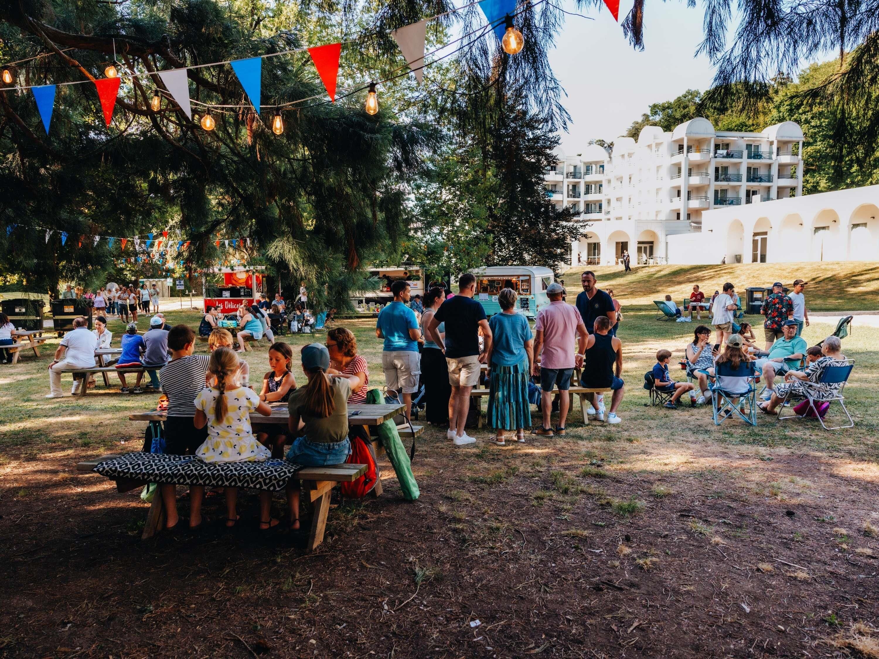 Eine gesellige Veranstaltung im Freien mit an Tischen sitzenden Personen, einigen Stehenden und einem Food-Truck im Hintergrund.