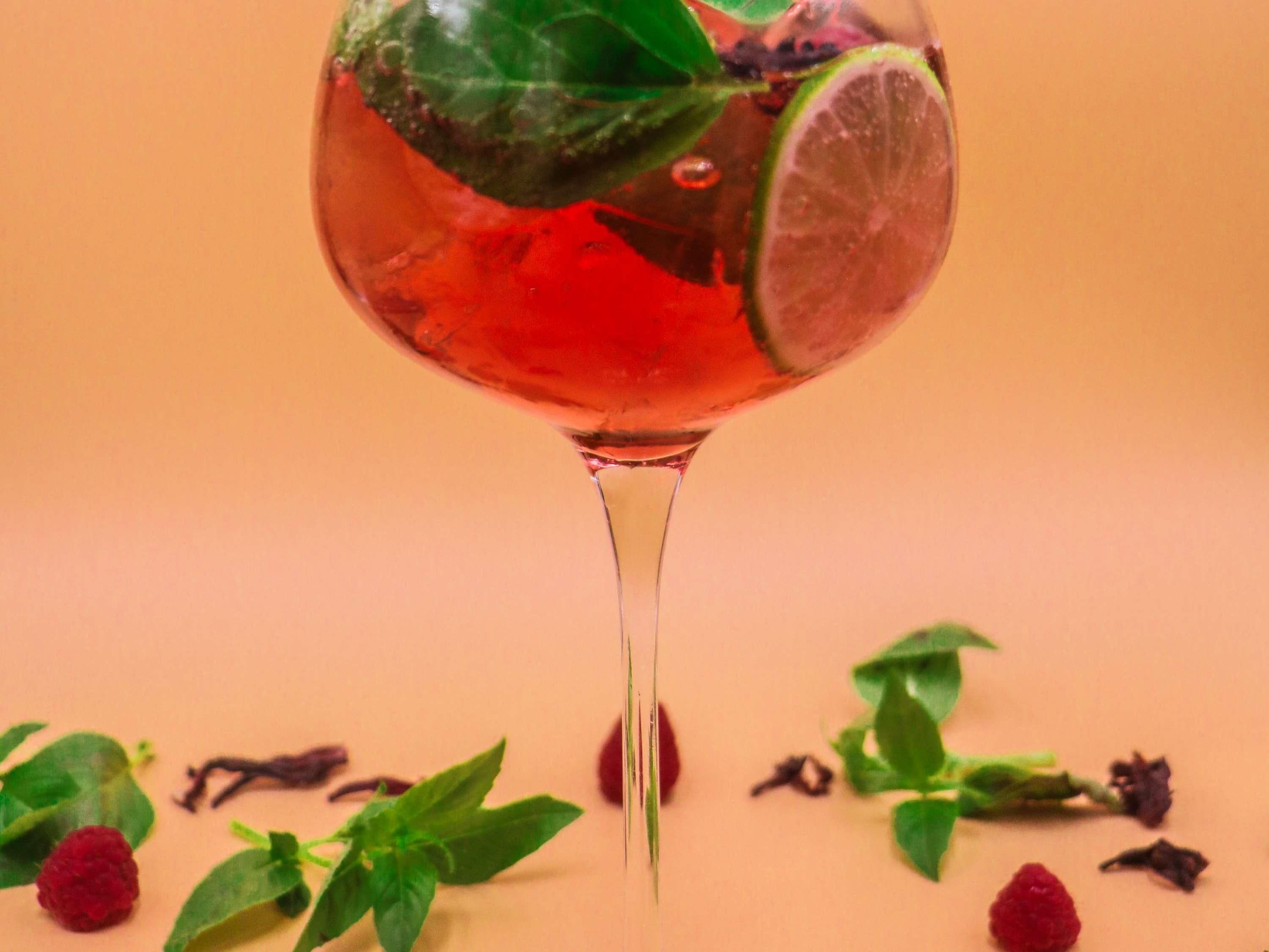 Un vaso de líquido rojo con lima y hojas de menta, rodeado de frambuesas y hojas de menta esparcidas sobre una superficie lisa.