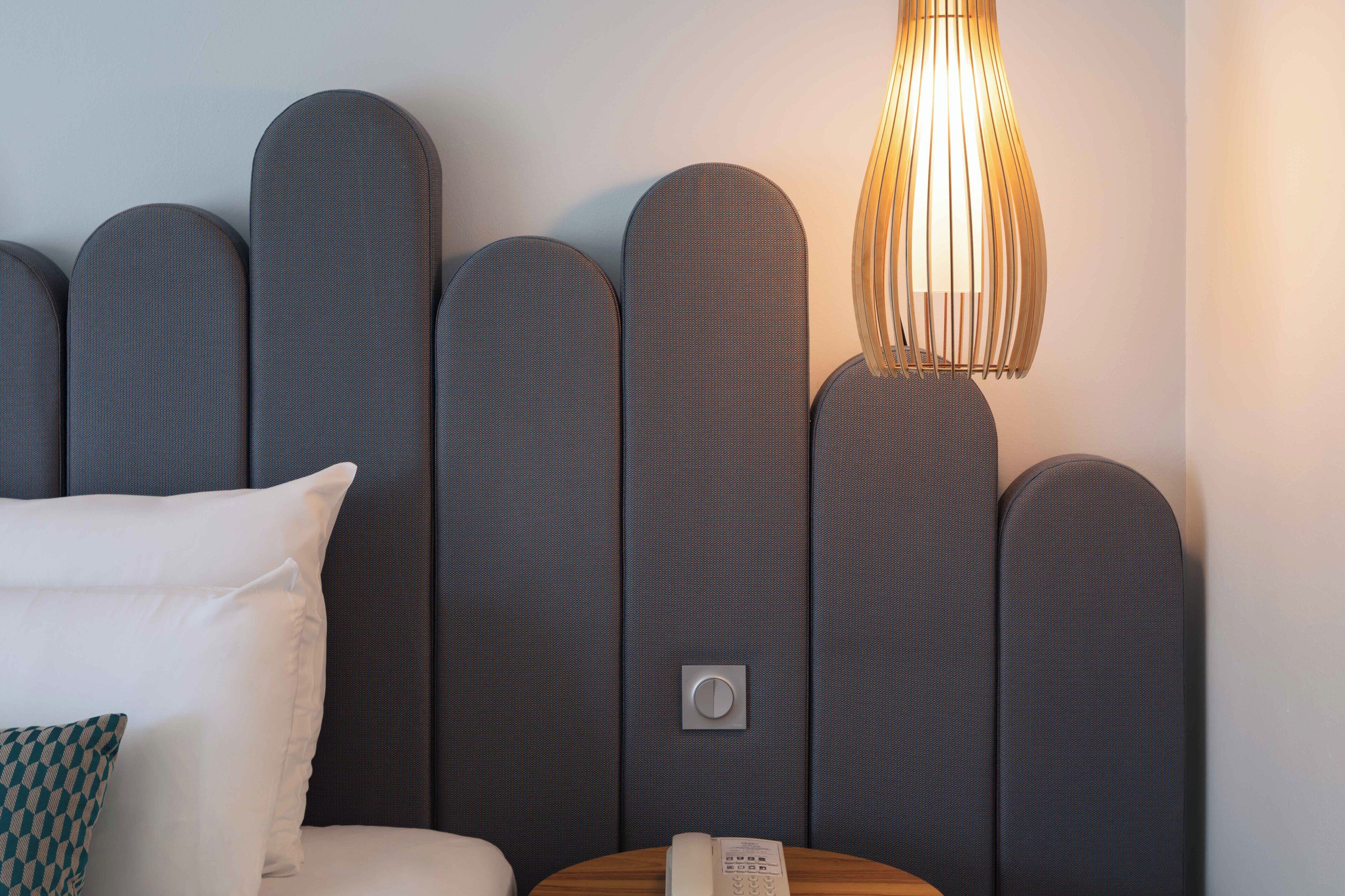 A modern hotel room with a headboard featuring a unique design and a bedside table with a lamp.