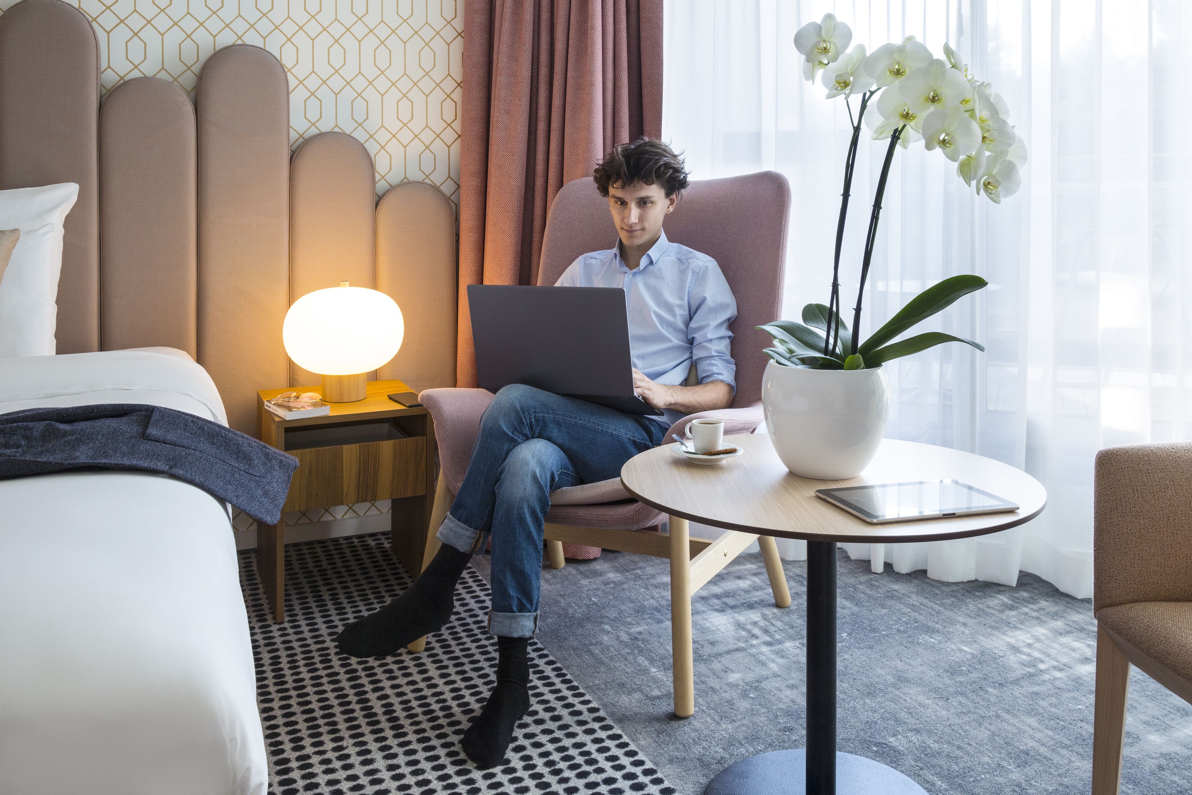 A hotel room with a bed, a chair, a laptop, a lamp, a potted orchid, and a tablet on a small table.