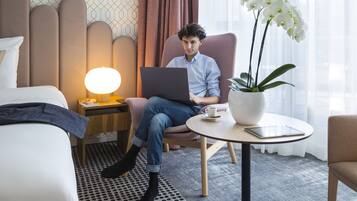 A hotel room with a bed, a chair, a laptop, a lamp, a potted orchid, and a tablet on a small table.