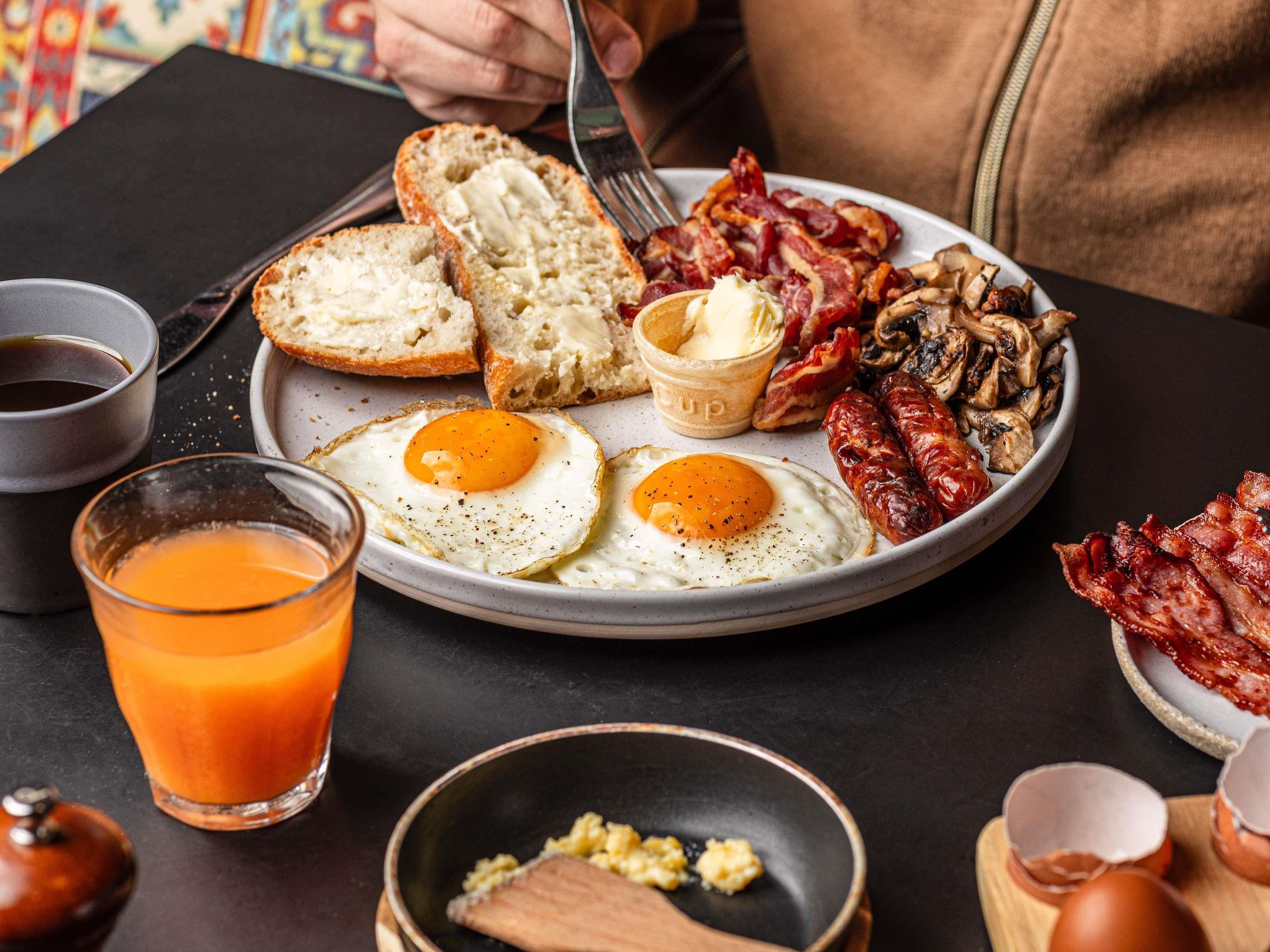 Un plato de desayuno con huevos, tocino, champiñones y tostadas, acompañado de un vaso de zumo de naranja y una taza de café.
