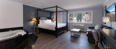 A modern hotel room with a four-poster bed, a separate bathroom with a bathtub, a small table, and a view of the outdoors through a window.