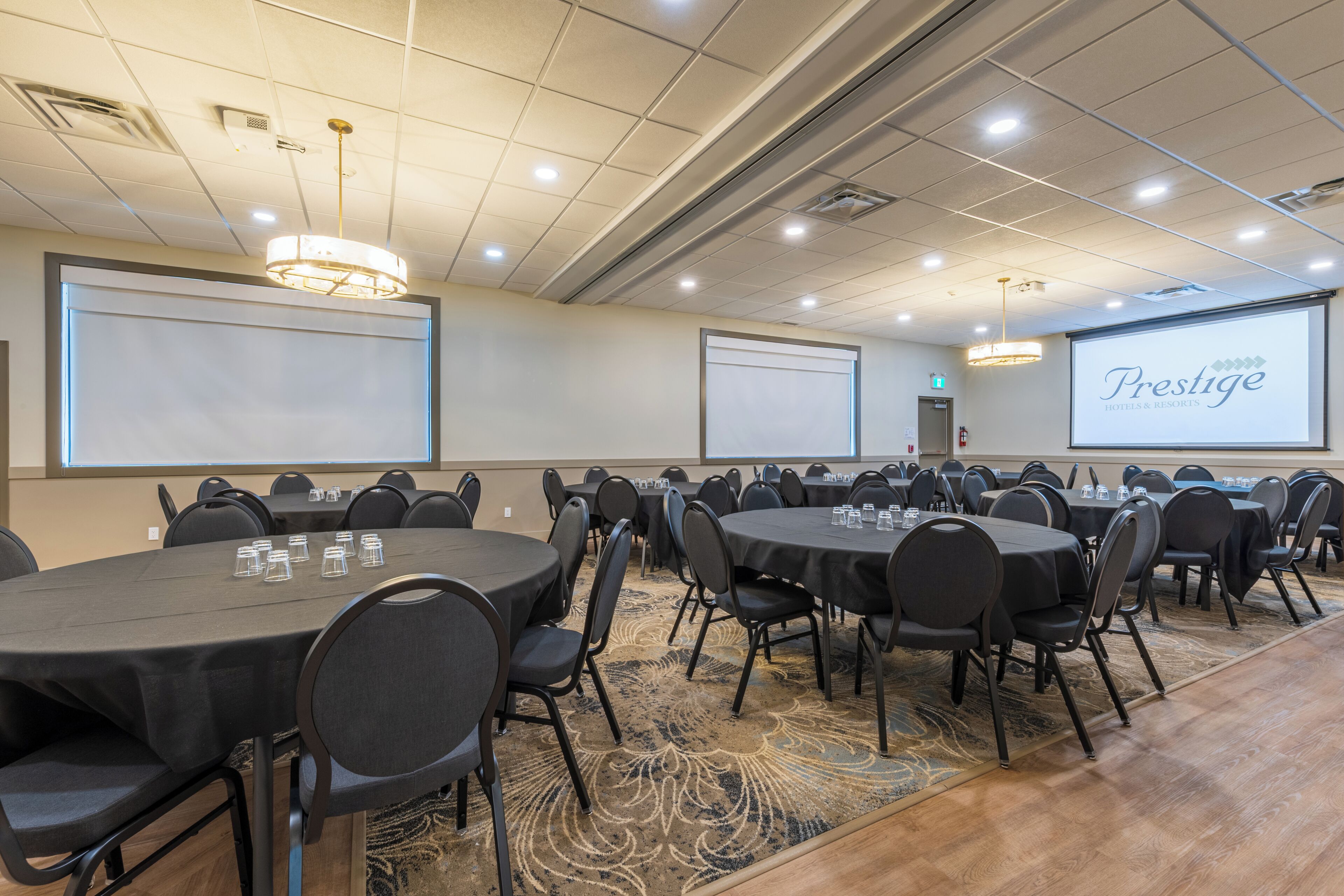 Une salle de conférence dotée de plusieurs tables et chaises, d’un écran de projection affichant le logo « Presgrave Hotels & Resorts », et d’un sol recouvert de moquette.
