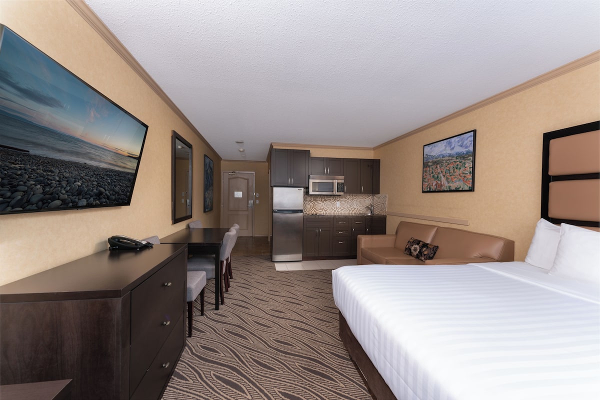 A hotel room with a bed, a desk, a chair, a kitchenette, and a painting on the wall.