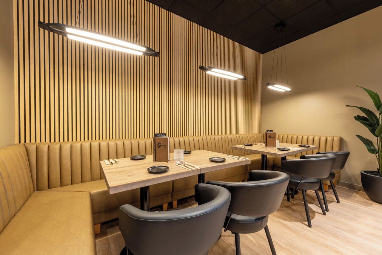 A modern restaurant with booth seating and rectangular tables, featuring wooden paneling on the walls and pendant lighting.