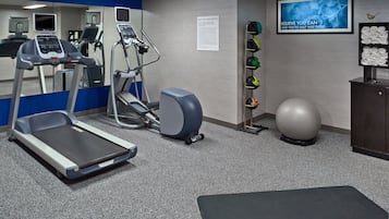 Un gimnasio con cintas de correr, elípticas, una bicicleta estática, un televisor montado en la pared, un estante con pelotas de ejercicio y una colchoneta.