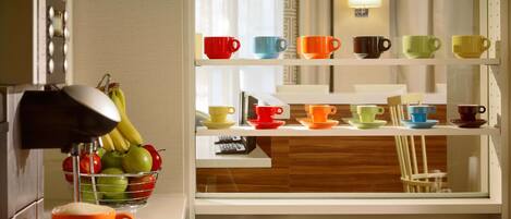 A kitchen with a coffee machine, fruit bowl, and a shelf with colorful cups and saucers.