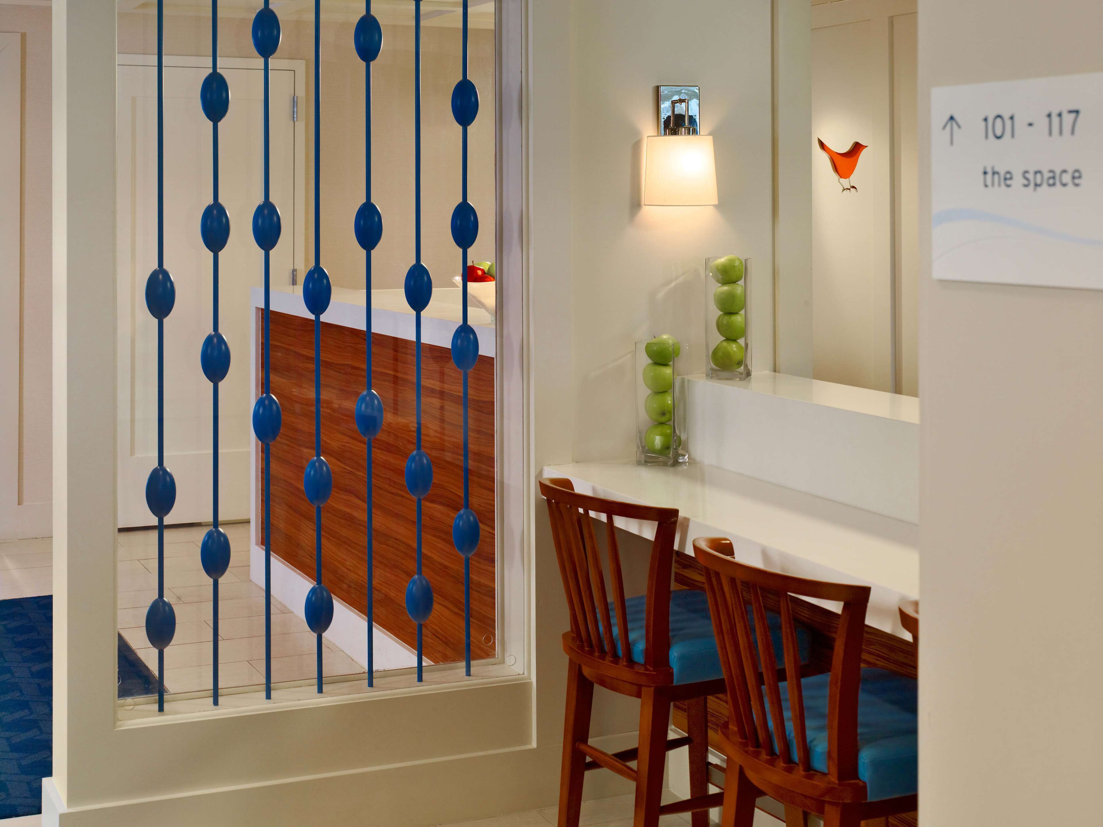 Ein Hotelzimmer mit Holzboden, einer Essecke mit weißem Tisch und Holzstühlen, einem blauen Dekorationselement am Fenster und einem Schild mit den Zimmernummern 101 bis 117.