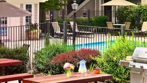 Eine Poolterrasse mit Holztischen und -bänken, umgeben von Grünpflanzen und einem Grill.