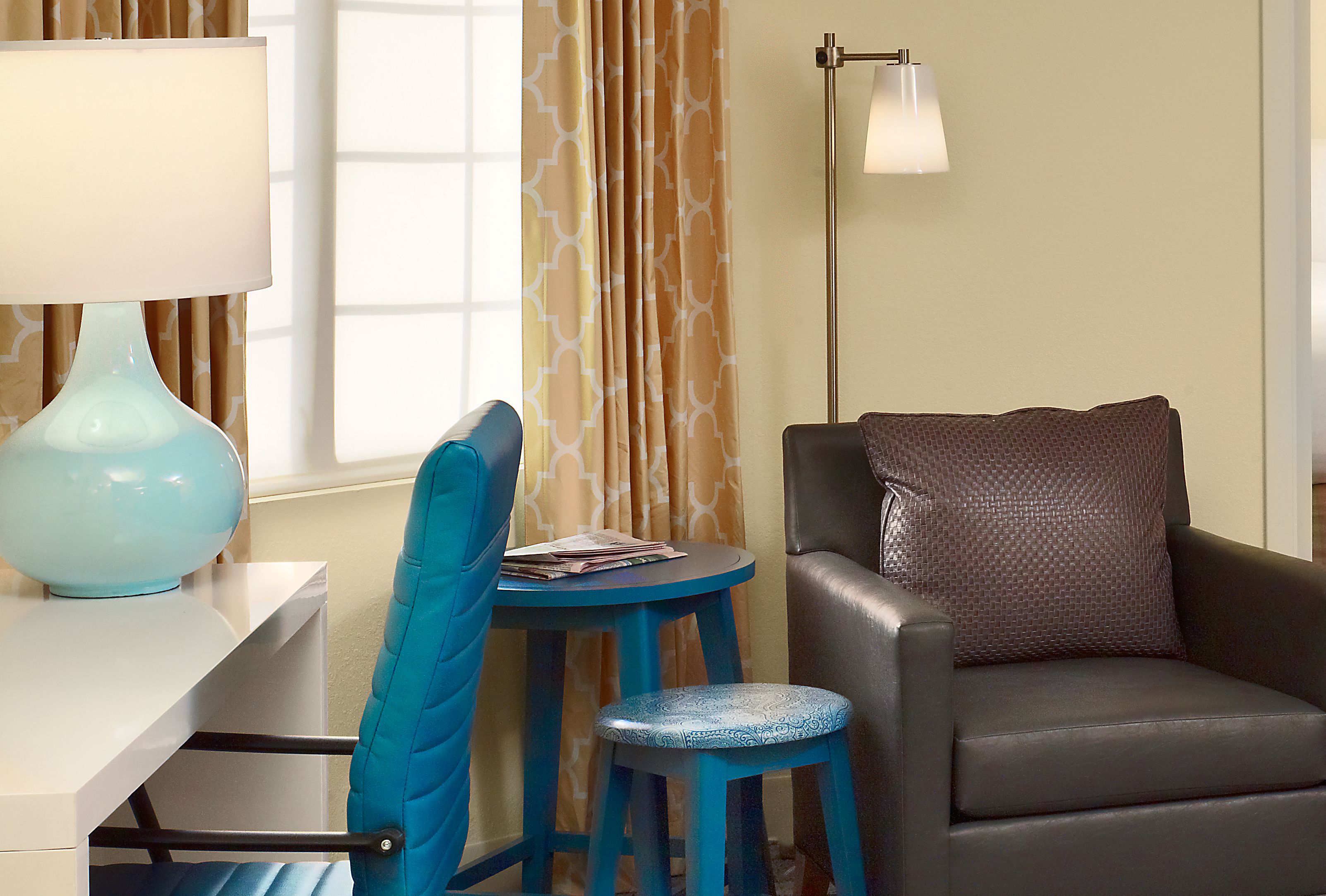 A room with a desk, a blue chair, a small stool, a sofa with a patterned pillow, and a floor lamp.